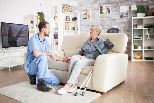 Male care taker in a nursing home holding arm of old woman sitting on the couch. Big tv in the background.