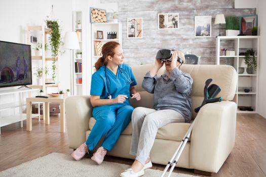 Amased elderly age woman while using virtual reality glasses in nursing home. Carevgiver and old woman on couch.