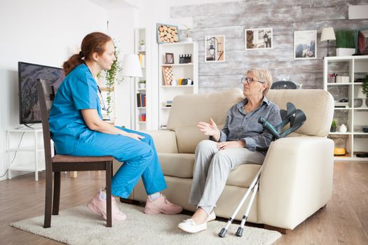 Nurse on a chair in a nursing home talking with elderly age woman with crutches.