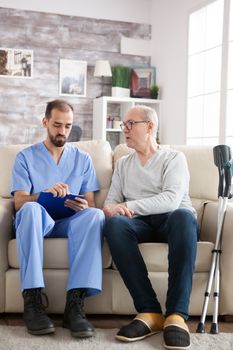 Male doctor with senior man sitting on couch in nursing home writing notes on clipboard. Old man with crutches.