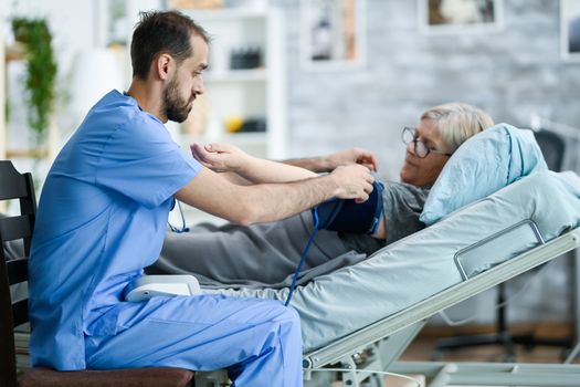 Male caretaker in a nursing home taking bood pressure of old woman laying in bed.