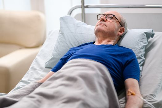 Thoughtful old man laying down on bed in a nursing home with transfusion on his arm.
