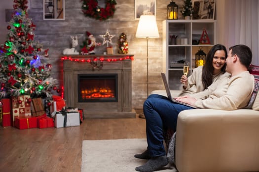 Beautiful couple smiling looking at each other celebrating christmas sitting on couch browsing on laptop. Woman holding a glass of champagne