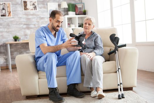 Young doctor sitting on couch in nursing home helping senior woman to use virtual reality glasses.