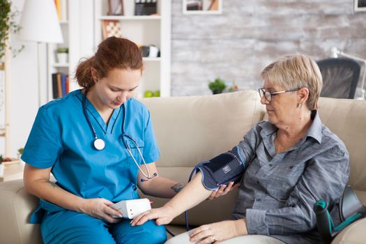 Nurse sitting on couch with old woman in nursing home checking her blood pressure with digital device.