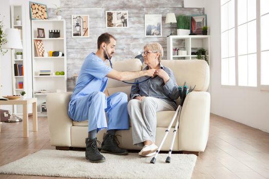 Doctor with stethoscope in a nursing home checking old woman heart beat sitting on couch. Old woman with crutches.