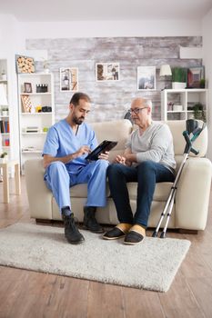 Young male doctor in nursing home taking notes on tablet pc while talking with senior man on the couch.