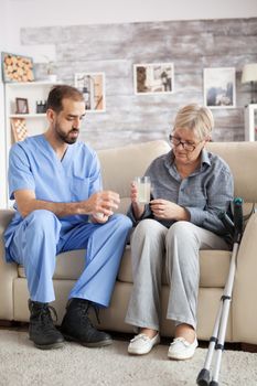 Male nurse sitting on couch with senior woman in nursing home helping her to take the medicine.