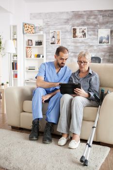 Young doctor in nursing home helping senior woman with crutches to use tablet computer.