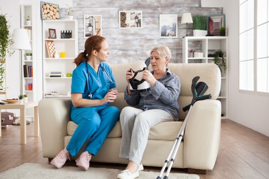 Elderly age woman sitting on couch with nurse holding virtual reality glasses in nursing home. Female caregiver.