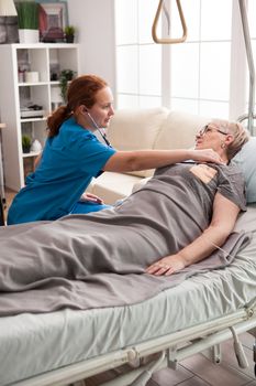 Female nurse using stethoscope to listen old woman heart in nursing home. Old woman laying in bed.