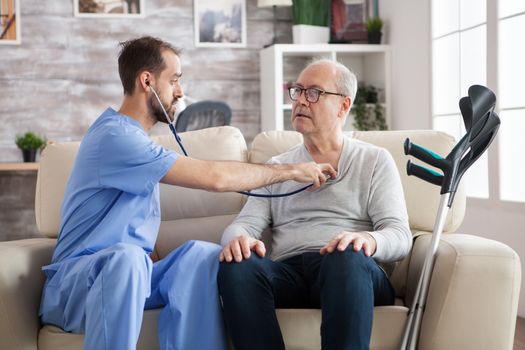 Young male doctor checking the heart of ill senior man in nursing home with stethoscope.