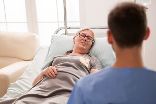 Old female laying on bed in a nursing home talking with male doctor sitting with the back at the camera.