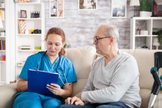 Female doctor with senior man sitting on couch in nursing home writing a prescription on clipboard.