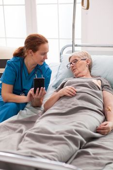 Female caretaker in nursing home talking with old woman sitting in bed and using mobile phone.