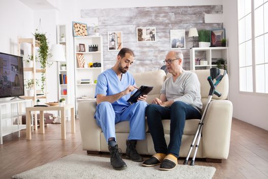 Young caretaker sitting on couch with senior man in nursing home using tablet computer.