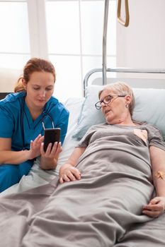 Female doctor helping old woman sitting on bed using mobile phone. Old woman in nursing home.