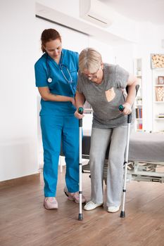 Old woman in nursing home walking with crutches with help from female nurse.