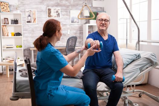 Female nurse helping old in with his rehabilitation in nursing home using dumbbells