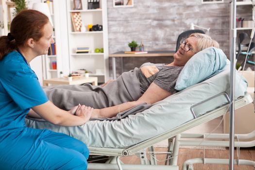 Smiling old woman laying on bed in nursing room holding nurse hand.