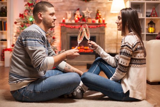 Romantic couple on christmas day in front of warm fireplace clinking a glass of champagne.
