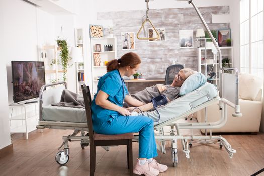 Young health caretaker taking blood pressure of old woman sitting on bed in nursing home.