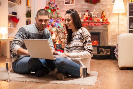 Relaxed couple on christmas day in front of fireplace smiling while using laptop.