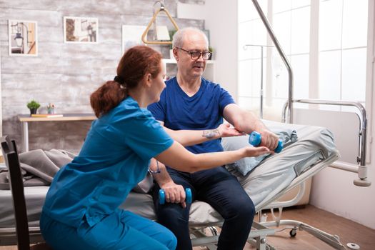 Senior man sitting on bed doing physiotherapy in nursing home using dumbbells with help from nurse.