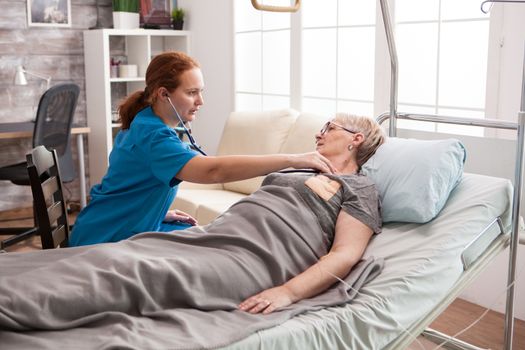 Young female doctor listening the heart beat of old woman laying in bed. Old woman in nursing home. Doctor with stethoscope.