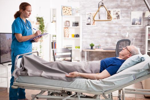 Female doctor taking notes on clipboard while talking with old patient in nursing home.