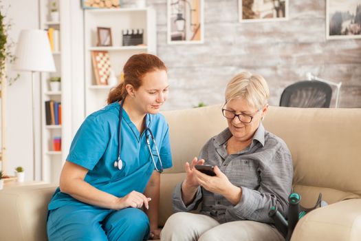 Female doctor in nursing home helping old woman to use her phone.