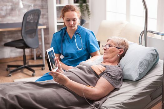 Old woman in nursing home sitting in bed using tablet computer the female nurse sitting next to bed.