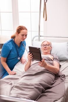 Senior woman lying in nursing home bed showing something to female nurse in tablet computer.