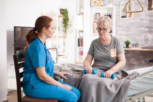 Female nurse helping old woman in nursing home with reheabilitation using dumbbells.