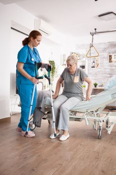 Caregiver in nursing home helping old woman to walk using crutches.