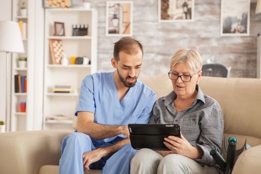 Male bearded doctor in nursing home helping senior woman to use her tablet computer.
