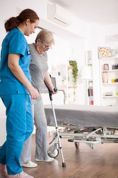 Female nurse helping old woman with walking problem in nursing home using crutches.