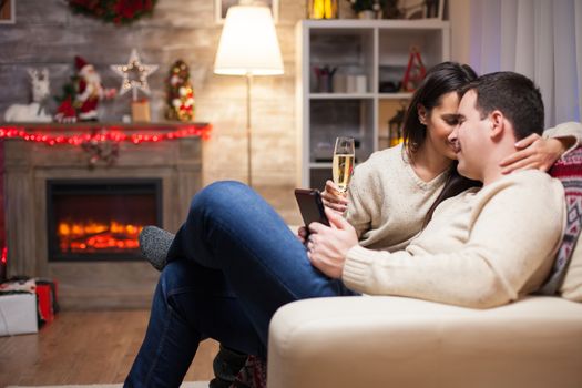 Young man holding his smartphone while showing affection to his wife on christmas day sitting on couch.