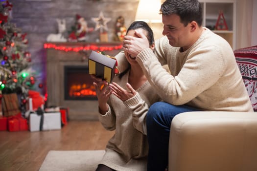 Young man covering woman's eyes while surprising her with gift on christmas day. Fireplace in living room.