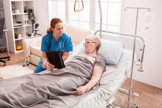 Female doctor in nursing home sitting next to senior woman lying in bed with her tablet computer.