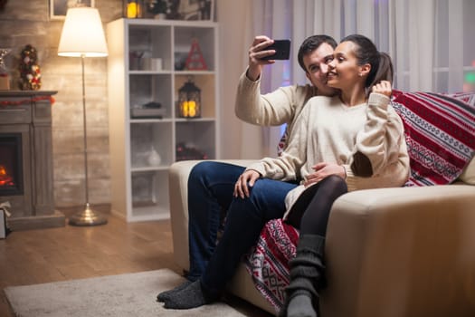 Smiling couple taking a selfie celebrating christmas day sitting on couch.