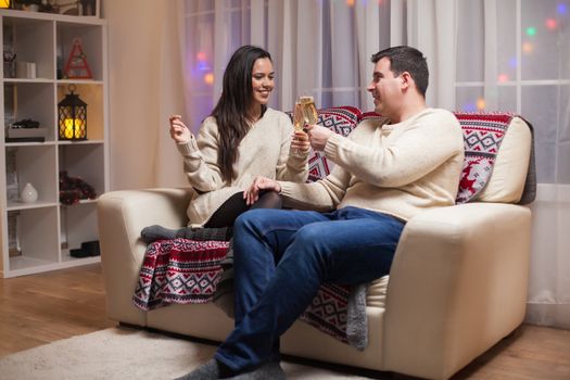 Beautiful young couple celebrating christmas sitting on couch clinking a glass of champagne.