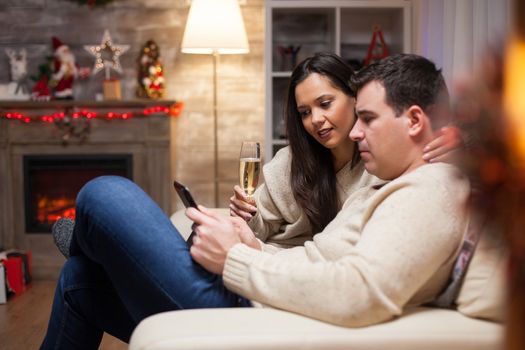 Beautiful wife holding a glass of champagne while her man is browsing on his smartphone on christmas day.