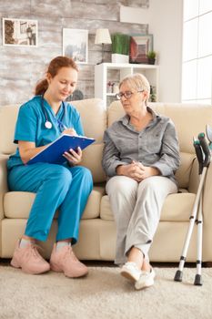 Young female doctor taking notes on clipboard while having a conversation with senior woman in nursing home.