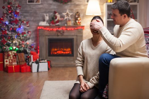 Young man covers his wife eyes on christmas day to give her a gift. Fireplace in the background.