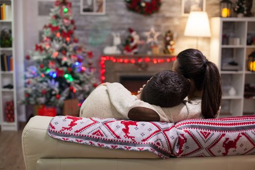 Young man resting his head on his girlfriend shoulder on christmas day sitting on couch. Back view.