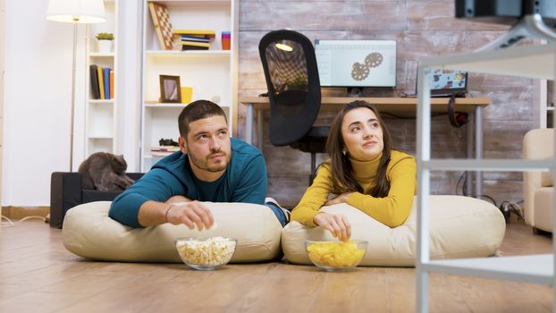 Cheerful couple watching tv sitting on pillows for the floor eating chips and popcorn with their cat in the background.