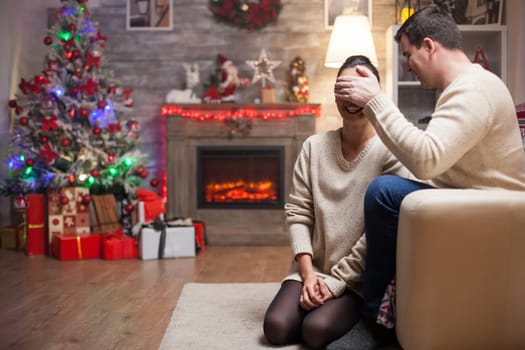 Woman with eyes covered on christmas day waiting to receive a present from her husband with fireplace in living room.