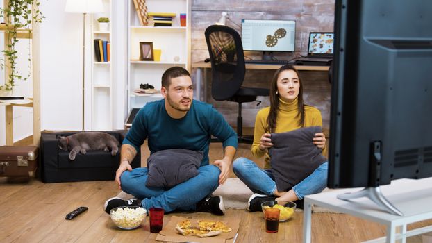 Caucasian young couple sitting on the floor in living room cheering up for their favorite sport team. Cat sleeping in the background.