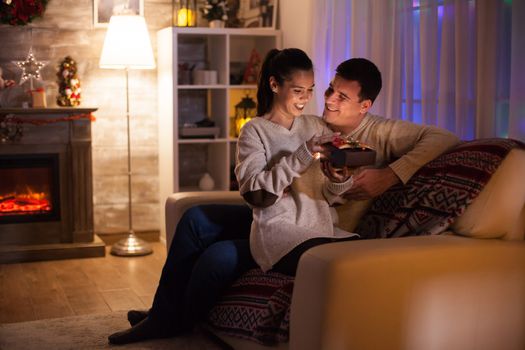 Young couple on christmas day sitting on couch in dark living room with fireplace in the background.